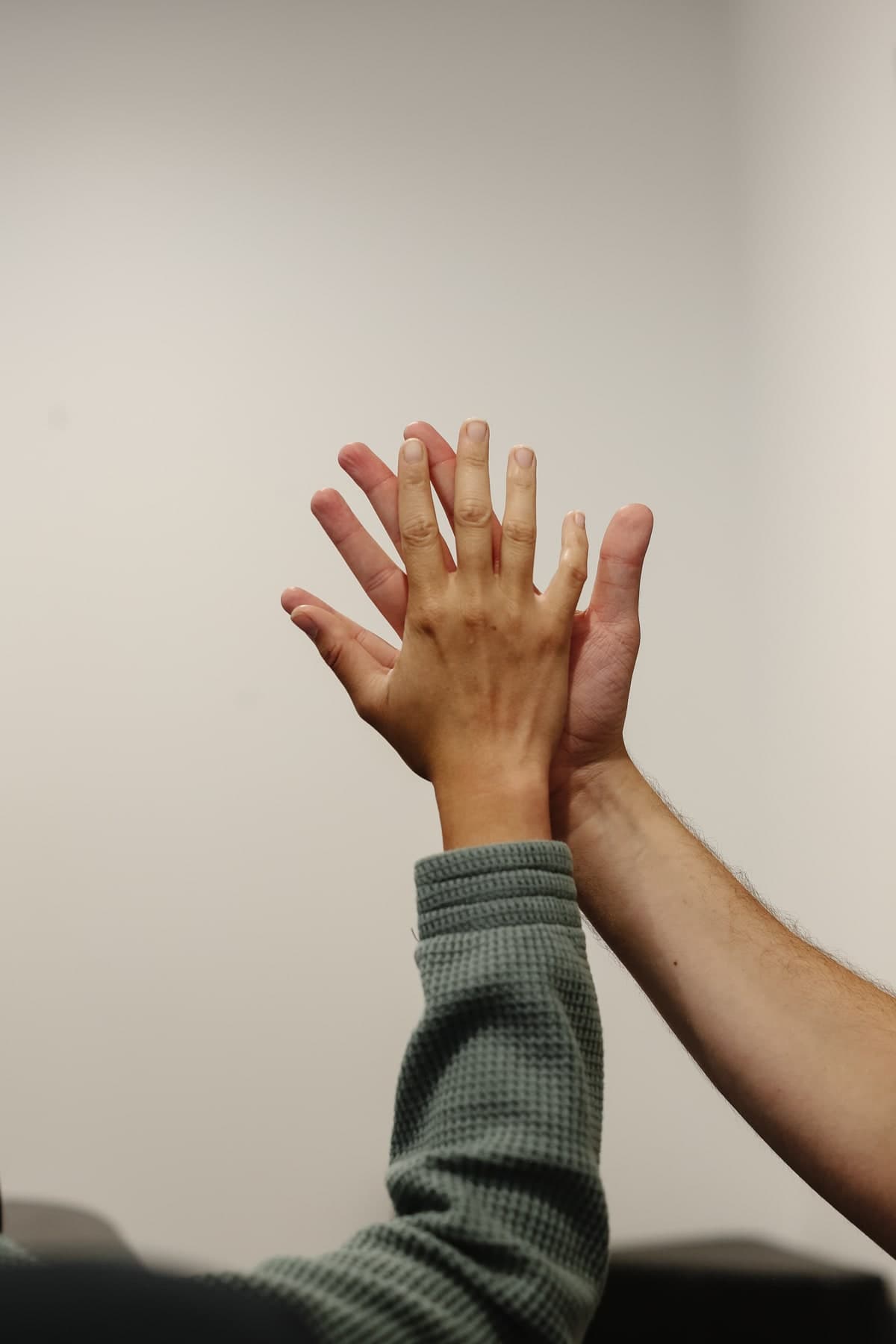 Two hands raised and meeting in a high five against a plain light grey wall, one wearing a grey waffle-knit sleeve